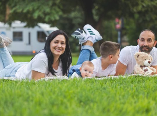 Happy family lying on grass with RV in the background, enjoying a sunny outdoor picnic.