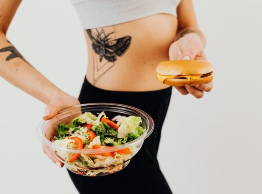 A woman with a tattoo holding a healthy vegetable salad and a cheeseburger, representing food choices.