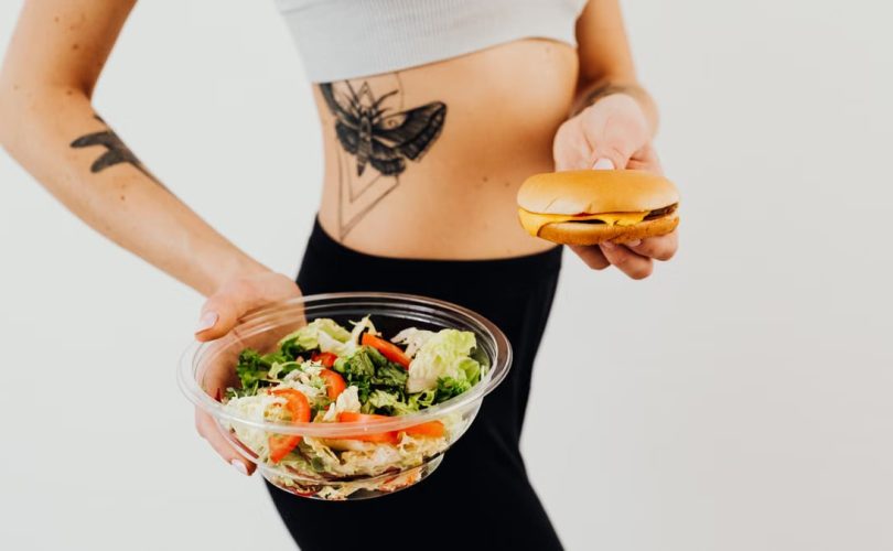 A woman with a tattoo holding a healthy vegetable salad and a cheeseburger, representing food choices.
